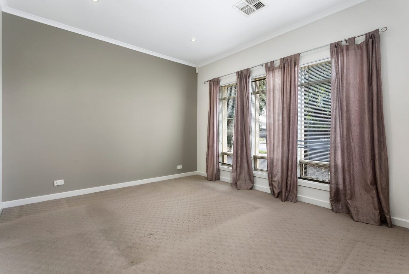 Large carpeted room with neutral walls, large windows, blinds, and elegant curtains.