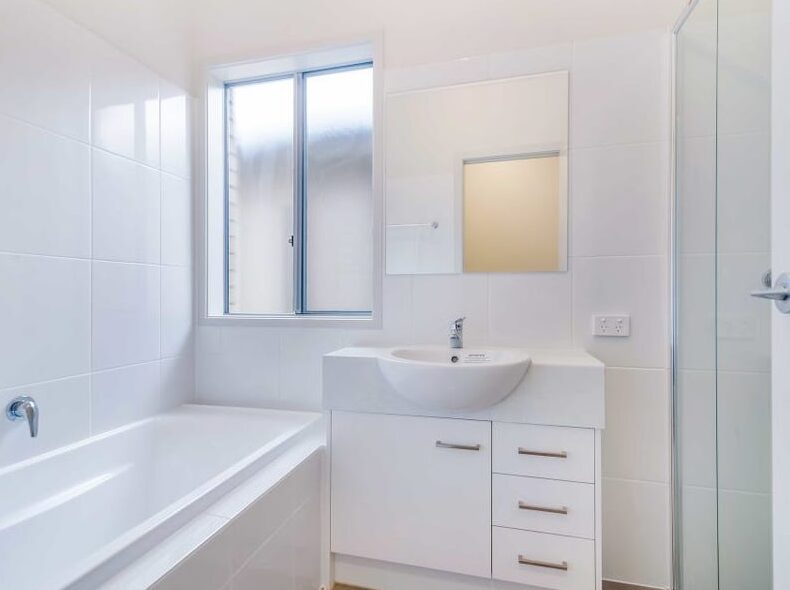 Bright white bathroom featuring a full-sized bathtub, single vanity sink, mirror, and glass shower enclosure.
