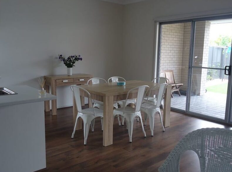Wooden dining table with white chairs set next to a sliding glass door leading outside.